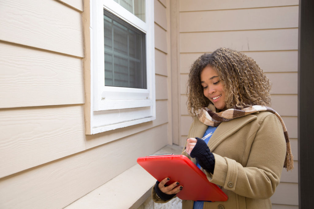 An image of a woman standing outside of a home, holding a red clip board.