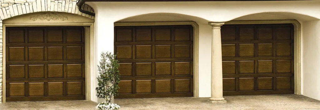 Raised panel wood garage doors. Three doors in medium dark brown with white pillars between.