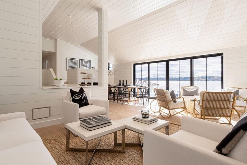 A photo of the inside of an open-concept living area in a home that has white shiplap walls, and black-framed lift and slide doors that show a view of a lake.