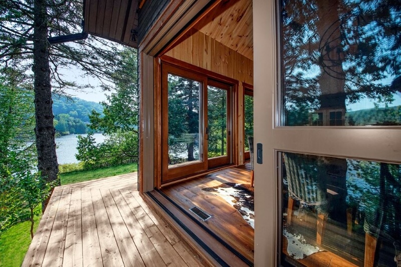 A photo of an open lift and slide door on a wooden home, leading out to a deck. The home is surrounded by trees and a lake.